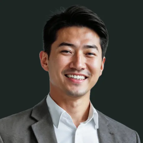 Smiling man with dark hair wearing a gray suit jacket and white shirt against a dark background.